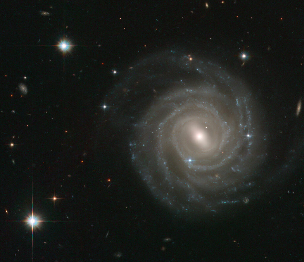 The galaxy captured in this image, called UGC 12158, certainly isn’t camera-shy: this spiral stunner is posing face-on to the NASA/ESA Hubble Space Telescope’s Advanced Camera for Surveys, revealing its structure in fine detail. UGC 12158 is an excellent example of a barred spiral galaxy in the Hubble sequence — a scheme used to categorise galaxies based on their shapes. Barred spirals, as the name suggest, feature spectacular swirling arms of stars that emanate from a bar-shaped centre. Such bar structures are common, being found in about two thirds of spiral galaxies, and are thought to act as funnels, guiding gas to their galactic centres where it accumulates to form newborn stars. These aren’t permanent structures: astronomers think that they slowly disperse over time, so that the galaxies eventually evolve into regular spirals. The appearance of a galaxy changes little over millions of years, but this image also contains a short-lived and brilliant interloper — the bright blue star just to the lower left of the centre of the galaxy is very different from the several foreground stars seen in the image. It is in fact a supernova inside UGC 12158 and much further away than the Milky Way stars in the field — at a distance of about 400 million light-years! This stellar explosion, called SN 2004ef, was first spotted by two British amateur astronomers in September 2004 and the Hubble data shown here form part of the follow-up observations. This picture was created from images taken with the Wide Field Channel of Hubble’s Advanced Camera for Surveys. Images through blue (F475W, coloured blue), yellow (F606W, coloured green) and red (F814W, coloured red) as well as a filter that isolates the light from glowing hydrogen (F658W, also coloured red) have been included. The exposure times were 1160 s, 700 s, 700 s and 1200 s respectively. The field of view is about 2.3 arcminutes across.