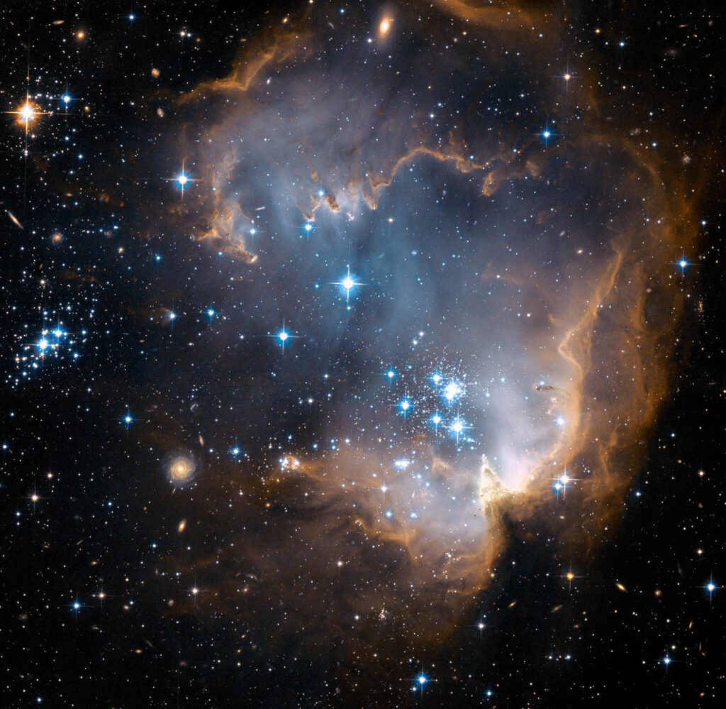 This image depicts bright blue newly formed stars that are blowing a cavity in the centre of a fascinating star-forming region known as N90. The high energy radiation blazing out from the hot young stars in N90 is eroding the outer portions of the nebula from the inside, as the diffuse outer reaches of the nebula prevent the energetic outflows from streaming away from the cluster directly. Because N90 is located far from the central body of the Small Magellanic Cloud, numerous background galaxies in this picture can be seen, delivering a grand backdrop for the stellar newcomers. The dust in the region gives these distant galaxies a reddish-brown tint.